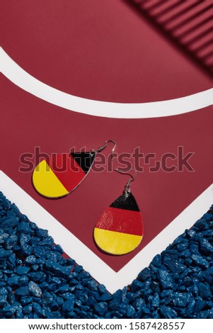 Object shot of leather pop-art earrings with German flag print, lying on a red surface with white stripes and blue stones on borders. 