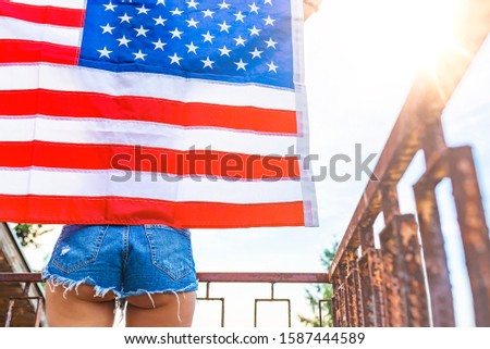 Woman holding American flag on blue sky background. Country, patriotism, independence day and people concept - happy smiling young woman in jeans with national american flag outdoors