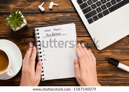 partial view of female hands near notebook with new years resolutions lettering near laptop, wireless earphones, plant and coffee cup on wooden desk