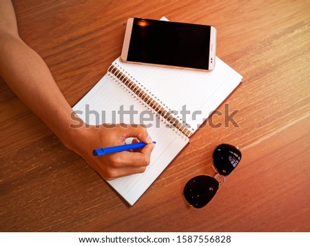 Top view of business accessories to short note on white note book which have the cellular phone or mobile phone or tablet and sun glasses on wooden table with sunshine lens flare.