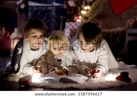 Three children, boy brothers, reading book at home at night on Christmas evening, lying on the floor