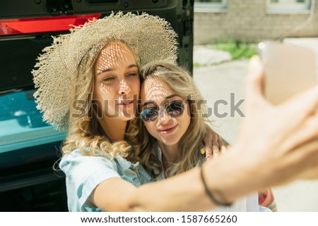 Young lesbian's couple preparing for vacation trip on the car in sunny day. Smiling and happy girls before going to sea or ocean. Concept of relationship, love, summer, weekend, honeymoon, vacation.