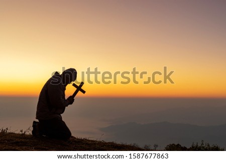 Silhouette human kneeling down praying and holding christian cross for worshipping God at sunset background.Christian, Christianity, Religion copy space background. 