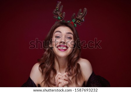 Indoor shot of young joyful lovely brunette with wavy hairstyle enjoying x-mas theme party masquerade and smiling happily to camera, dressed in elegant clothes over claret background