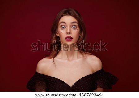 Open-eyed young pretty female with brown wavy hair wearing festive evening makeup while standing over claret background, rounding amazedly eyes and looking at camera with shocked face