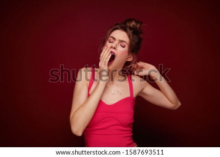 Sleepy young attractive brown haired female in festive clothes covering her mouth with raised palm and yawning with closed eyes, coming home after party with friends, isolated over burgundy background