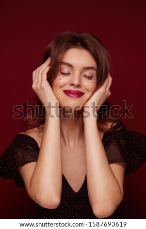 Pleased attractive young brown haired female lady with festive makeup wearing elegant black top with red dots while posing over claret background, smiling nicely with closed eyes