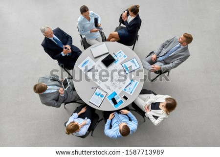 people, work and corporate concept - business team with gdgets and papers at round office table Royalty-Free Stock Photo #1587713989