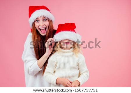 ,happy redhead ginger woman and cute little blonde girl hiding eyes under santa claus hat and having fun together in studio pink background