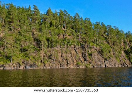 Blue river and blue clear sky. Along the shores are green pine forest and yellow birch trees. The rocky left bank is reflected in the water.