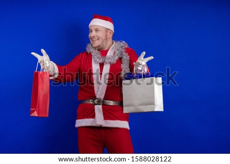 Emotional male actor in a costume of Santa Claus holds gift bags with purchases in his hands and poses on a blue background