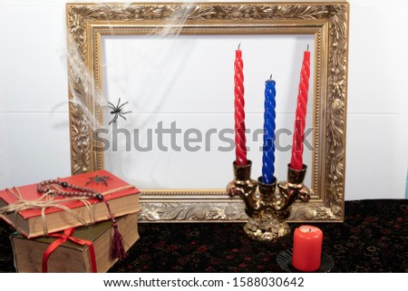 On a velvet tablecloth is a large red candle and a candlestick with candles. Nearby are old shabby books with a rosary and a spider on it. A cobweb with a spider hangs on a picture frame.