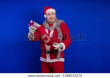 Emotional male actor in a costume of Santa Claus holds in his hands a Christmas sock and a box with gifts and poses on a blue background