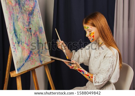 The young girl of the artist in a light white dress, paints a picture on canvas in the workshop. The face is stained with paints. A young student uses brushes, canvases and easels. Creative work