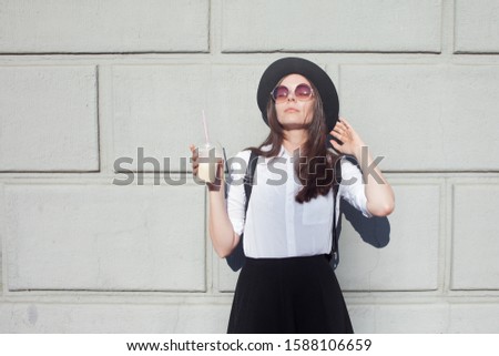Young woman in hat and round sunglasses walking in city. Girl tourist enjoys the walk. Hipster in a hat with a backpack, Drinking coffee smoothies. On wall background, copy space