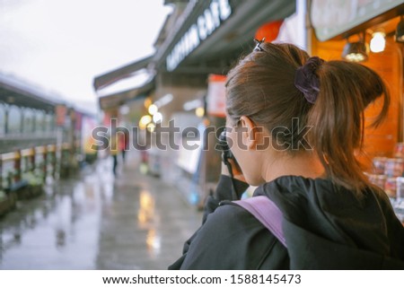 Back view image of woman traveling alone taking photo with camera in her hands at Shifen train station in Taiwan during rainy day, head shot of woman on journey and blurry background of train station