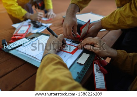 Construction miners worker writing his name and sign onto personnel red danger tag and placing locking into isolation permit safety control lockbox while his friends locking on at the background Royalty-Free Stock Photo #1588226209