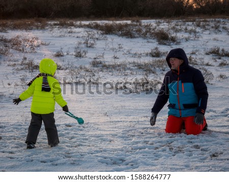 Dad in bright ski suit stands with charming boy in yellow jacket holds device for sculpting snowballs from snow. child and father on background of beautiful winter landscape sunset. village Holidays 