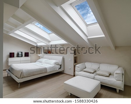 interior shot of a bedroom in the modern attic-room in the foreground the leather sofas with footrest in the background there is the bed illuminated by two skylights Royalty-Free Stock Photo #1588330426