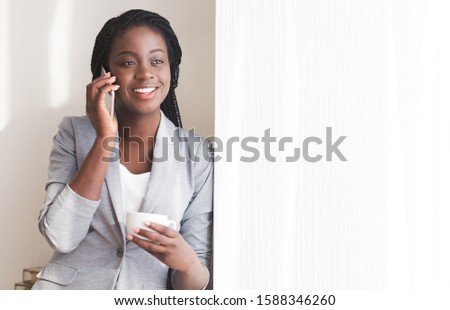 Coffee break. Cheerful young black businesswoman talking on cellphone and drinking coffee near window in office, copy space