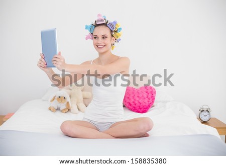 Cheerful natural brown haired woman in hair curlers taking picture of herself with a tablet in bright bedroom