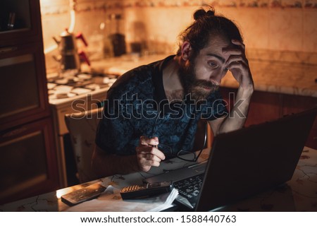 Hipster young man checking bills in the kitchen and using a laptop. Financial problems.