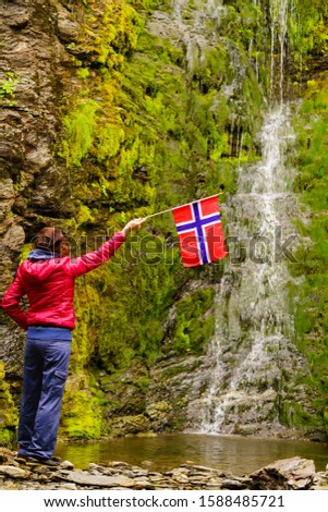 Tourist woman with norwegian flag enjoying waterfall view in green summer mountains, Norway.