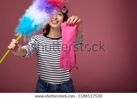 A smiling girl shows in the frame a dust brush and a pink rubber glove for cleaning. Lovely girl cleans, brings cleanliness. Photo on a pink background the girl in the curlers cleans.