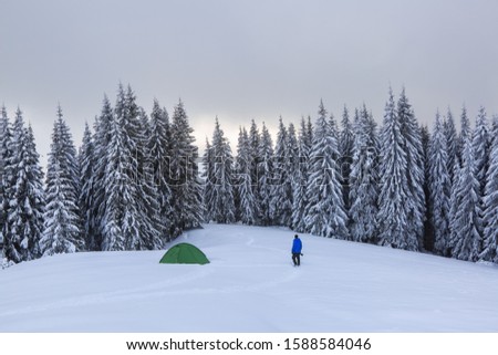 Mountain winter landscape. Tourist stands on the path next to the green tent. Wild forest. Touristic camping rest place. Location Carpathian, Ukraine, Europe. Snowy wallpaper background.
