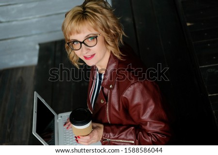 Adult girl with glasses sitting with a laptop and drinks coffee in a paper cup. Business woman working at a computer in a cafe.