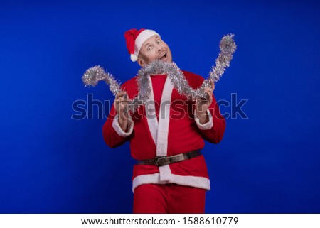 Male actor in a suit and hat of Santa Claus with a silver tinsel garland dancing and posing on a blue background