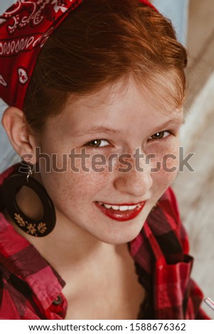 Cute red-haired girl with freckles, dressed in a red bandana and a plaid shirt on the background of the kitchen. Advertising, pin-up photo concept