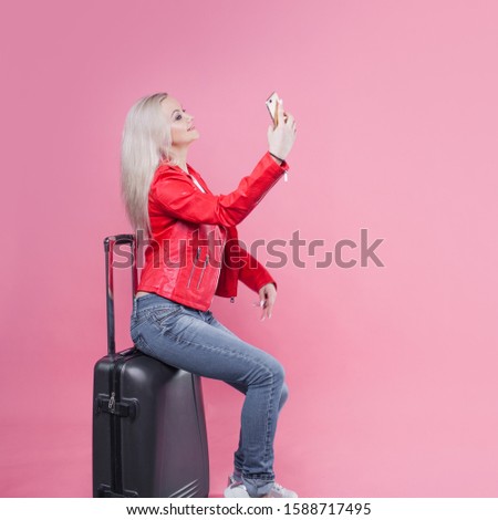 young female tourist in a red jacket and with Luggage on a pink background. Tourist girl ready to fly. Charming blonde sits on a suitcase and takes a selfie