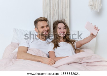Happy and dreamy young adult woman with man spending morning in bedroom, lying on bed in nightwear, making self portrait on modern smartphone