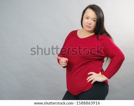 Adult plus size woman pointing with finger at copy space with funny face expression, on grey