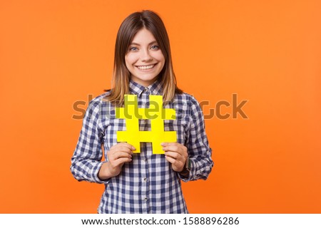 Viral media tag. Portrait of young happy brunette woman with charming smile wearing checkered shirt standing holding large big yellow hashtag sign. indoor studio shot isolated on orange background