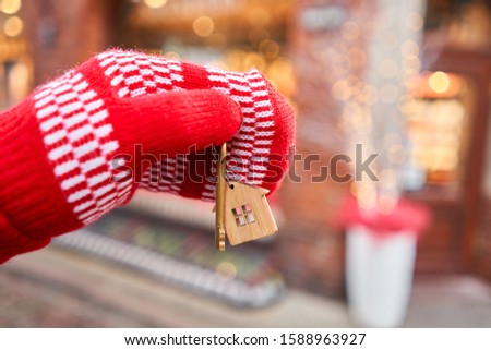 Mortgage or rent concept. Hand in red mitten holding key with house shaped keychain. Real estate, hypothec, moving home or renting property. Christmas mood in blurred background.