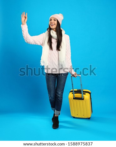 Young woman in warm clothes with suitcase on blue background. Winter vacation