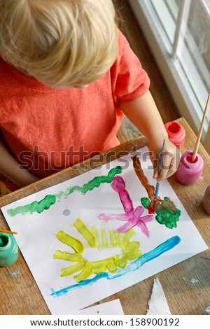 a creative young child can be seen from above painting a happy picture of sun, grass and trees on a piece of paper