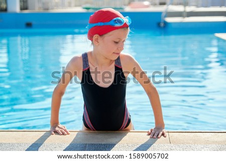 Beautiful smiling girl in outdoor swimming pool, child in swimming cap, swimsuit and goggles. Sunny summer day, sports healthy lifestyle for children.