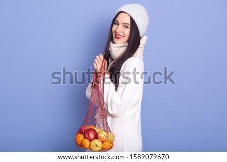 Horizontal picture of charming magnetic young woman holding apples and tangerines in net handbag, smiling sincerely, being satisfied, wearing warm items of clothes. New Year atmosphere concept.