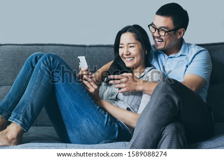 using cell phone couple asian lover happy enjoy sitting on sofa at home looking on mobile laugh and smiling together search information from internet.concept for technology contact communication