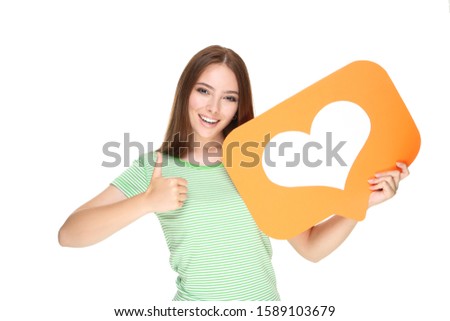 Young woman holding paper card with heart and showing thumb up on white background
