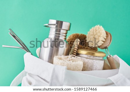 Reusable plastic free items in cotton shopping bag. Glass jar, metal straws, aluminum bottle and wooden cleaning brush on green background