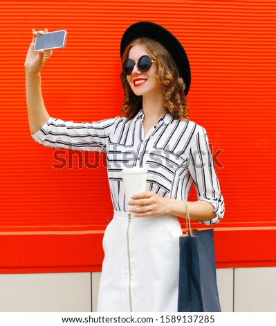 Beautiful young smiling woman taking selfie picture by smartphone with coffee cup and shopping bags wearing white striped shirt, black round hat on city street over red wall background