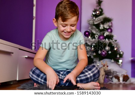 Little  cute blond laughing boy    sitting   in pijamas at  the floor at home  near a christmas three  playing  video games on tablet  