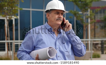 Mature male landscape architect (60 years old) wearing hardhat and carrying plans talks on mobile phone. Many baby boomers continue working into their senior years.                               