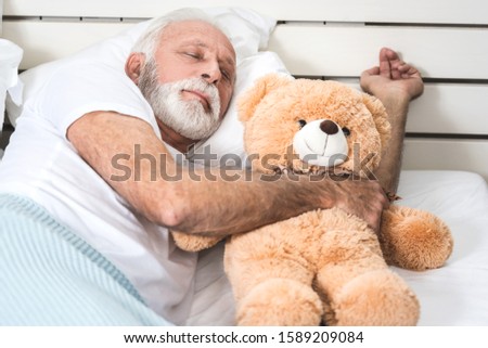 Old man with white hair and beard sleeps alone with teddy bear in his bedroom.  Closeup picture