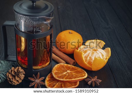 Winter Christmas vintage still life: a French press with tea, tangerines, dried oranges, cinnamon sticks and anise stars on a textured, black, wooden background.