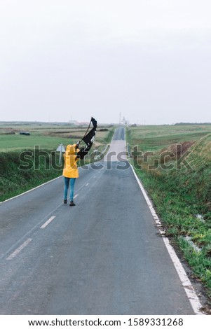 A young girl with a yellow raincoat walks along a road between fields heading to the coast waving a pirate flag.